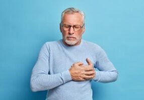 Senior man in spectacles presses hand to chest has heart attack suffers from unbearable pain closes eyes wears optical glasses poses against blue background. People age and problems with health